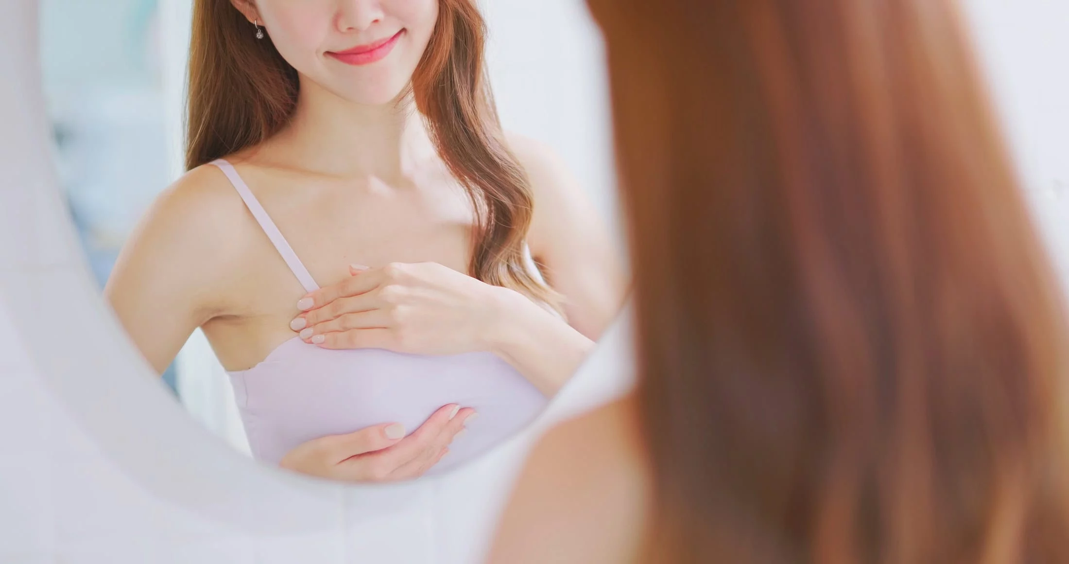 身穿白色背心的女子在鏡子前檢視自己的乳房，欣賞著魔滴隆乳所達到的美胸效果。