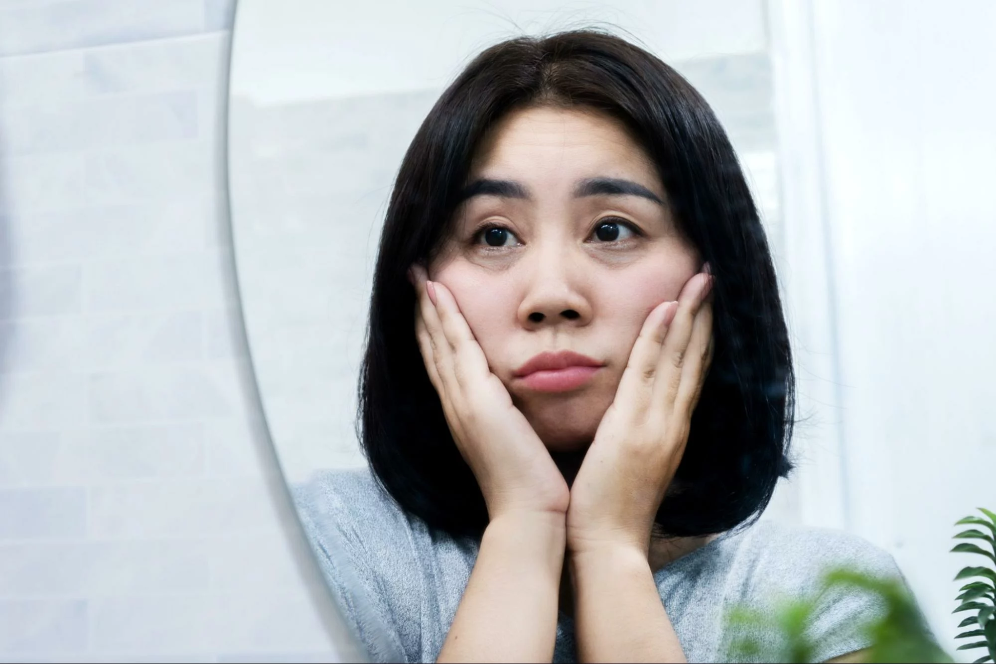 一位女子凝視著鏡子中的自己，一邊將臉輕輕地託在手中，一邊思考著如何消除眼袋，重拾電眼雙眸。